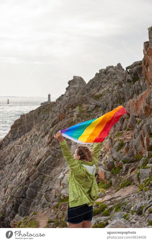 Blonde Frau hebt ihre Arme an der Spitze des Umhangs und schwenkt eine Regenbogenflagge, im Hintergrund eine Felswand und ein Leuchtturm im Meer. lgbtq