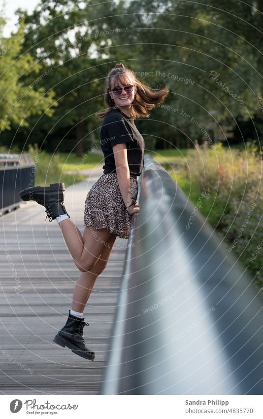 Junge Frau auf einer Brücke lächelt in die Kamera Brückengeländer Jung Frau Stiefel Rock stylisch langhaarig Sonnenbrille feminin Außenaufnahme schön