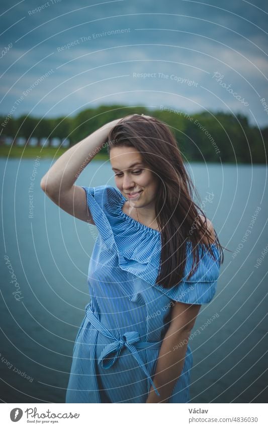 Nettes brünettes Modell mit langen glatten dunklen Haaren in einem blauen Sommerkleid mit einem schönen realistischen Lächeln posiert an einem See in der Tschechischen Republik. Europäische Frau. Ungezwungenes Porträt mit realistischem Lächeln