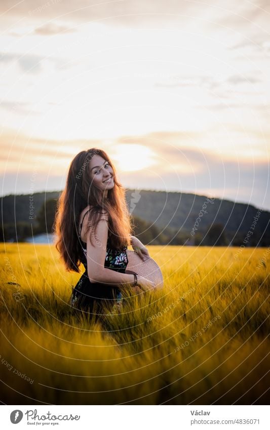 Schönes realistisches Lächeln einer Brünetten im schwarzen Kleid, die in der Mitte eines Feldes bei Sonnenuntergang steht. Unverfälschtes Porträt der inneren Emotionen. Kräuseln Sie Ihr Haar