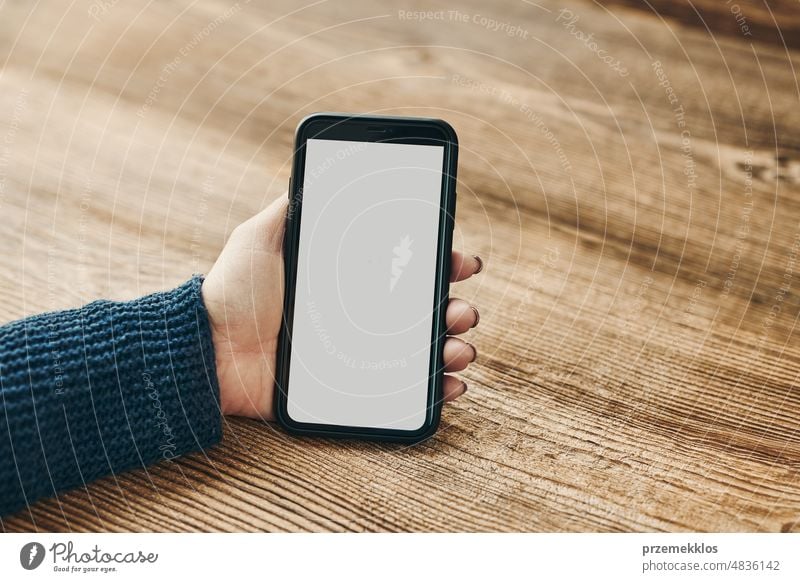 Weibliche Hand hält Smartphone Mockup mit fester weißer Farbe leeren leeren Bildschirm auf hölzernen Hintergrund. Person mit Technologie. Mobiltelefon mit Kopie Raum links
