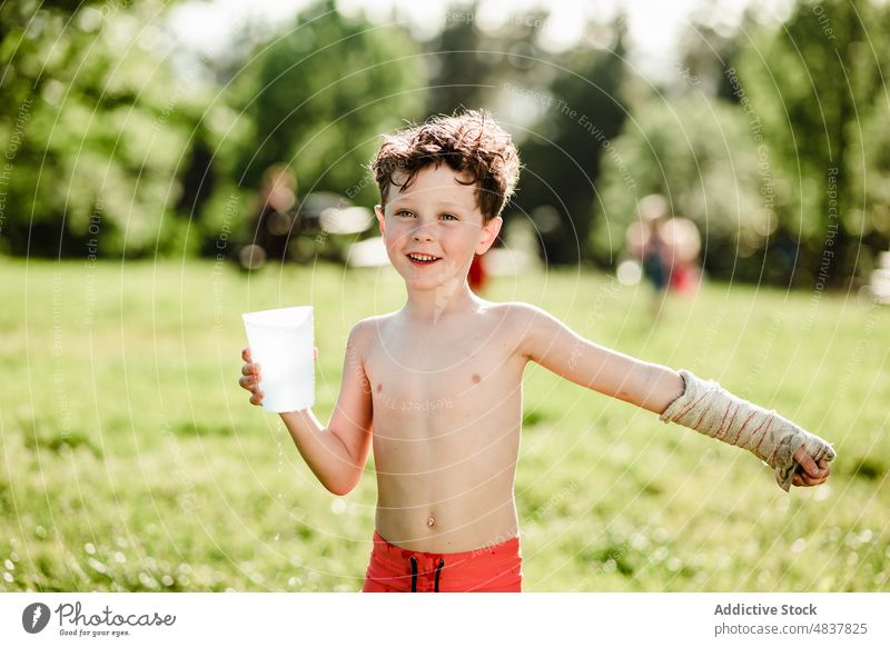 Glücklicher Junge mit Glas Wasser Sommer ohne Hemd platschen heiter Eimer Kindheit positiv Rasen Garten Freude Lächeln Feiertag Natur sorgenfrei genießen