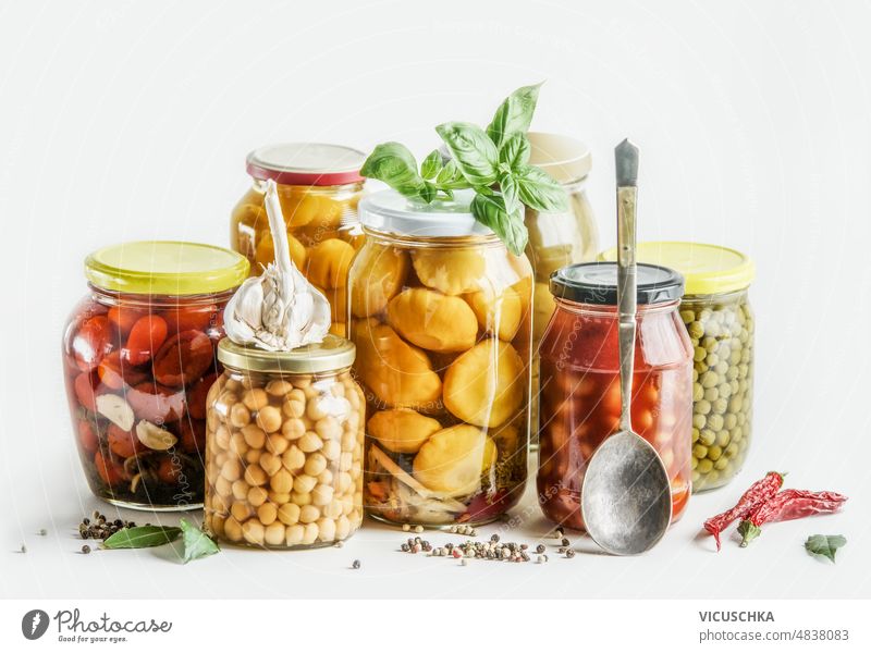 Eingemachtes Gemüse in Gläsern mit Kräutern, Gewürzen und Löffel auf weißem Hintergrund: eingemachte Kichererbsen, Zucchini, Bohnen und Tomaten. konserviert