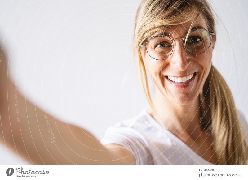 Attraktive blonde Frau lässig gekleidet und Brille schauen lächelnd breit. Schöne Frau mit fröhlichen Ausdruck beim Posieren gegen weißen Hintergrund, Stretching Arm zur Kamera für ein Selfie