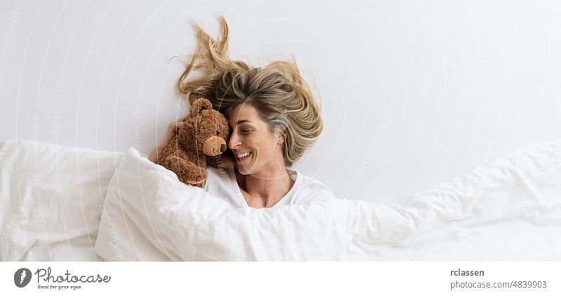 Frau, die mit ihrem Teddybär kuschelt, der auf einem weißen Bett schläft und unter einer Decke liegt, Ansicht von oben, Platz für Ihren individuellen Text. Top