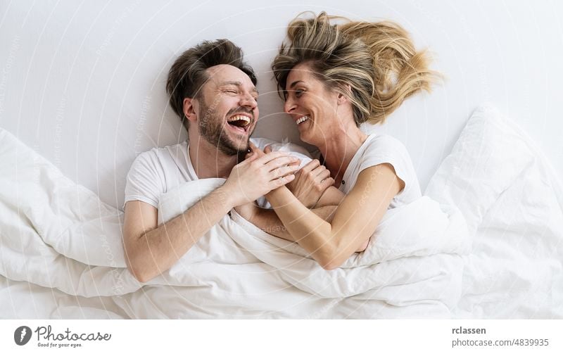 Draufsicht auf ein junges Liebespaar, das lachend und lächelnd auf einem Bett mit Decke liegt. Sie schauen sich gegenseitig mit Vertrauen in der Liebe an Paar