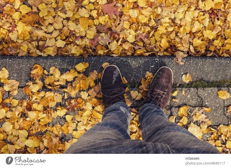 Junger Mann steht an einem Bordstein mit Laubblättern im Herbst auf der Straße, persönliche Perspektive von oben. pov Menschen Lifestyle Punkt Ansicht Weg