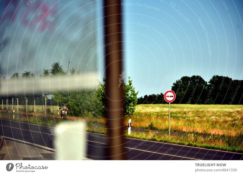Blick aus Bus Haltestelle über Landstraße mit Verkehrszeichen Überholverbot und Begrenzungspfosten auf ein sommerliches Feld mit Wald im Hintergrund und wolkenlosem Himmel; eine Radfahrerin scheint sich zu nähern