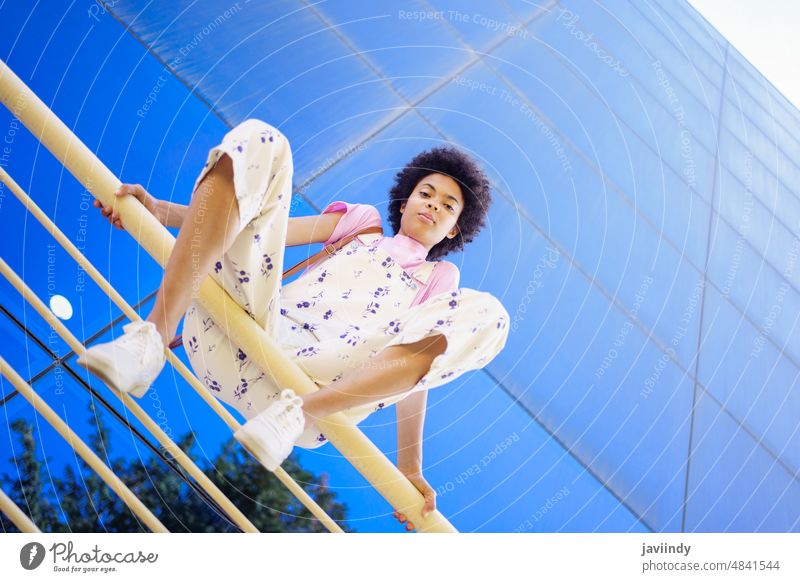 Afroamerikanische Frau in Freizeitkleidung, die auf einem Geländer eines modernen Gebäudes sitzt. positiv in die Kamera schauen Fröhlichkeit Afro-Look Frisur