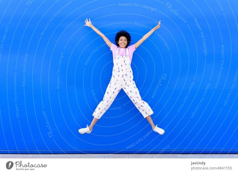 Afroamerikanische Frau, die mit erhobenen Armen gegen eine blaue Stadtmauer springt. springen Fröhlichkeit Freude Glück Lächeln Afro-Look Frisur schwarz lockig