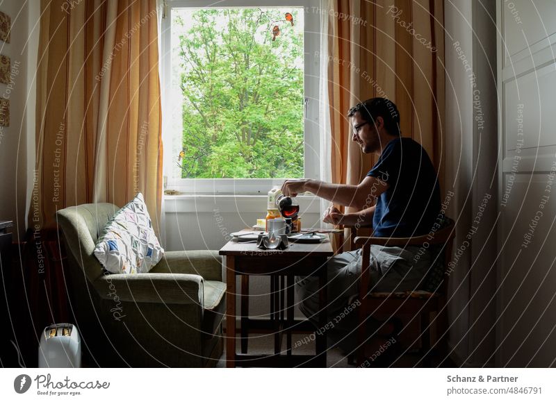 Mann sitzt an einem Fenster und frühstückt alleine zuhause Frühstück essen Sessel gemütlich entspannt einsam Vorhang Aussicht Person Zuhause Quarantäne