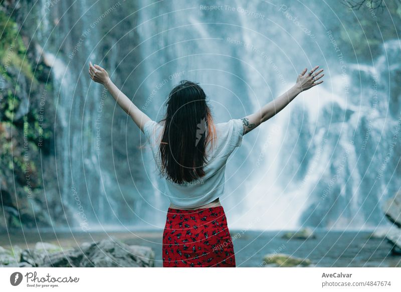 Junge Frau springt, feiert und drückt ihr Glück vor einem Wasserfall aus, reist und tourt alleine. Freiheit in der Natur, unabhängiger Lebensstil und Freiheitskonzept. Bild zurück
