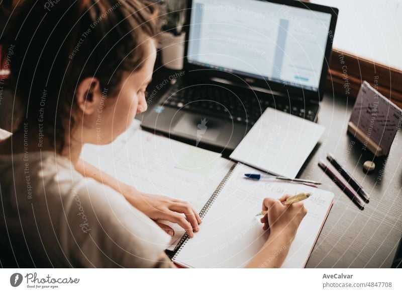 Draufsicht auf den Rücken einer jungen Frau, die zu Hause am Schreibtisch studiert und arbeitet, Hausaufgaben macht, sich mit dem Lehrbuch auf die Prüfung vorbereitet und Notizen macht, selektiver Fokus auf den Bleistift.