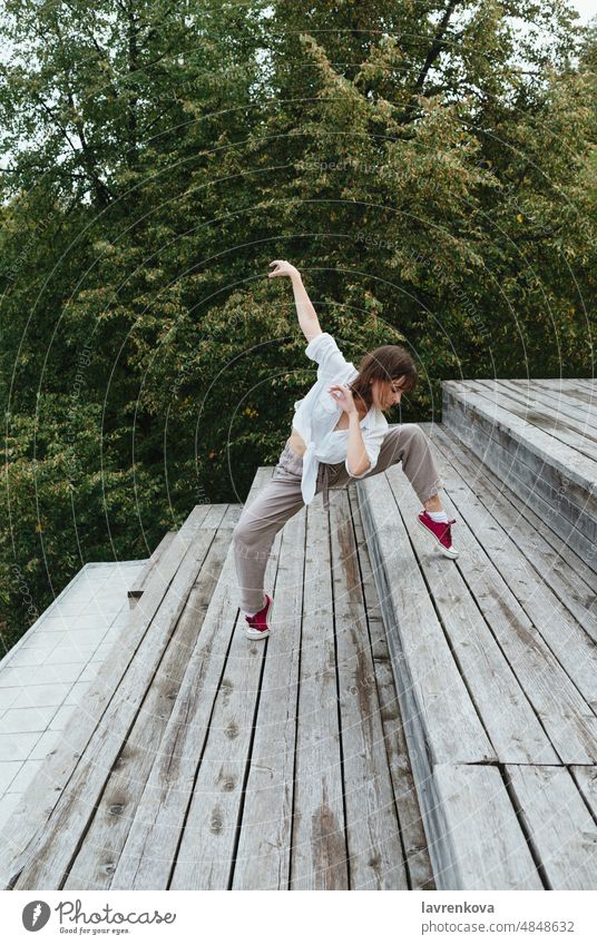 Junge Frau in weißem Hemd und grauer Leinenhose tanzt auf der Treppe vor dem Laubwerk, Tänzer Choreograph im Freien Mädchen stylisch urban Bewegung Sport