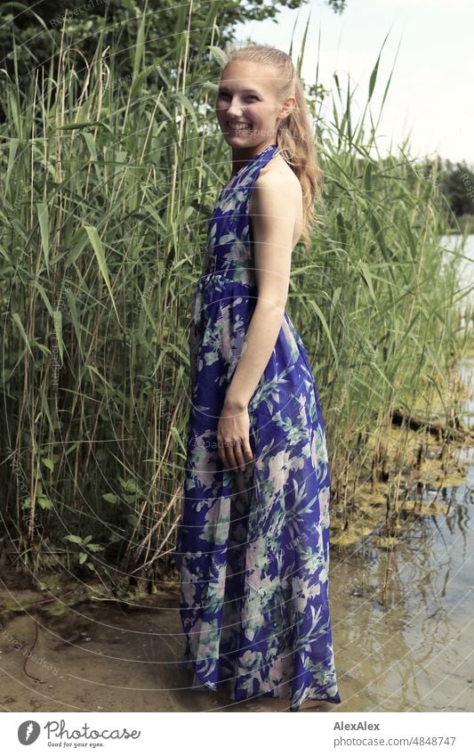 Junge, rotblonde, langhaarige Frau steht im Kleid mit dem Füßen im See am Ufer und lächelt seitlich in die Kamera jung schön schlank draussen Gras Natur