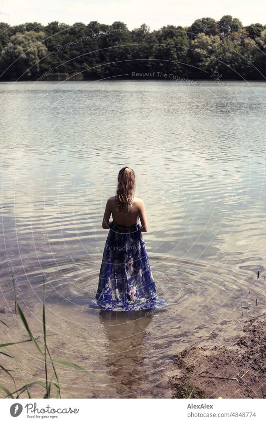 Junge, rotblonde, langhaarige Frau steht im Kleid mit dem Füßen im See am Ufer mit dem Rücken zur Kamera jung schön schlank draussen Gras Natur fröhlich