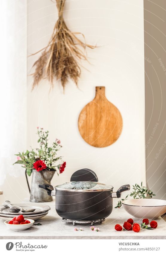 Kochtopf mit Frühstücksbrei auf Küchentisch mit Beeren Haferbrei Platten Blumen Schüsseln Wand Hintergrund Schneidebrett vorbereitend gesunde Ernährung
