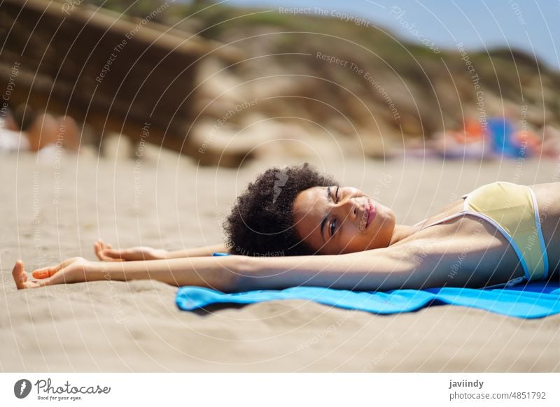 Zufriedene junge afroamerikanische Frau liegt am Sandstrand und zwinkert der Kamera zu Strand Sonnenbad sich[Akk] entspannen Arme hochgezogen Urlaub Meeresufer