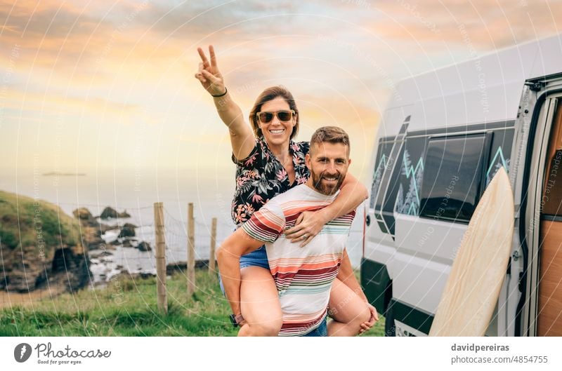 Ein Paar, das sich mit der Kamera vergnügt und neben dem Wohnmobil Huckepack nimmt jung Glück Spaß schauende Kamera Ausflug Siegeszeichen v-Zeichen Hand Lächeln