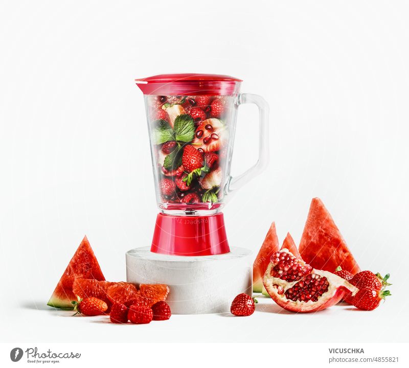 Glasmixer gefüllt mit roten Früchten: Wassermelonenscheiben, Granatapfelkerne, Himbeeren, Erdbeeren und Grapefruit auf Produktpodest vor weißem Hintergrund mit Zutaten.