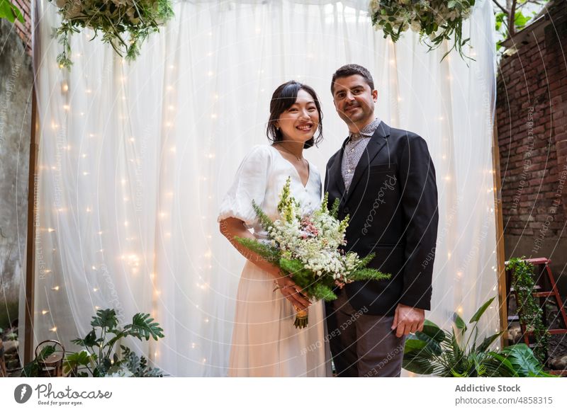 Neuvermähltes multiethnisches Paar mit Blumenstrauß Jungvermählter Braut striegeln Umarmung Blütezeit Hochzeit Bogen rassenübergreifend vielfältig asiatisch