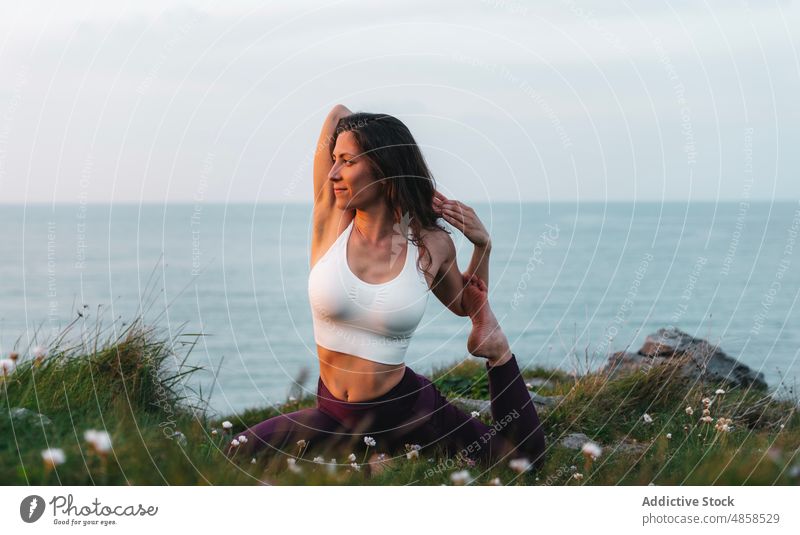 Eine fitte Frau macht Yoga am Meeresufer Athlet Sportbekleidung grasbewachsen eka pada raja kapotasana üben Natur Gesunder Lebensstil Hügel Wasser Seeküste