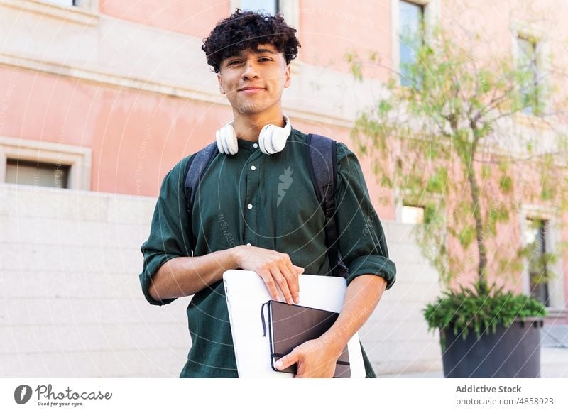 Lächelnder ethnischer Mann mit Laptop auf der Straße Schüler Netbook Kopfhörer Großstadt Gebäude Sommer Lifestyle Glück Freude heiter sorgenfrei Notizblock