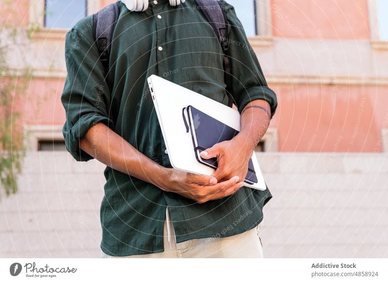 Anonymer ethnischer Mann mit Laptop auf der Straße Schüler Netbook Kopfhörer Großstadt Gebäude Sommer Lifestyle Freude heiter sorgenfrei Notizblock Planer