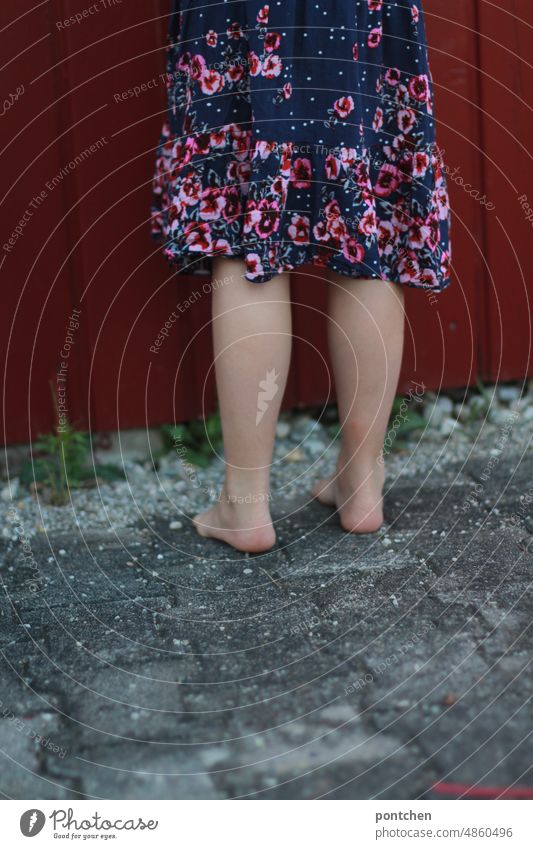 Kinderspiel. Verstecken Unterkörper. Abgewandt Kind in Kleid steht vor einer roten bretterhütte, hütte. stehen kleid abgewandt wegdrehen spielen barfuß