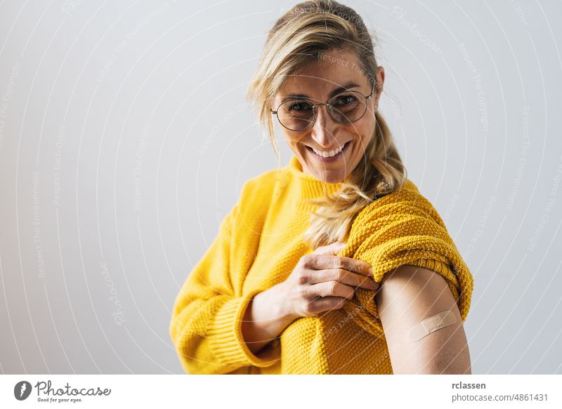Porträt einer reifen Frau mit Brille, die lächelt, nachdem sie eine Corona-Impfung erhalten hat. Frau hält ihren Hemdsärmel hoch und zeigt ihren Arm mit Verband, nachdem sie geimpft wurde.
