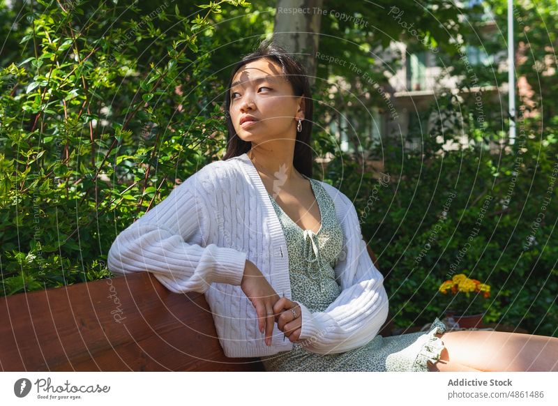 Ruhige asiatische Frau sitzt auf einer Bank im Park Vorschein Straße charmant verträumt Sommer Windstille Sonnenlicht feminin Stil Großstadt Dame ethnisch