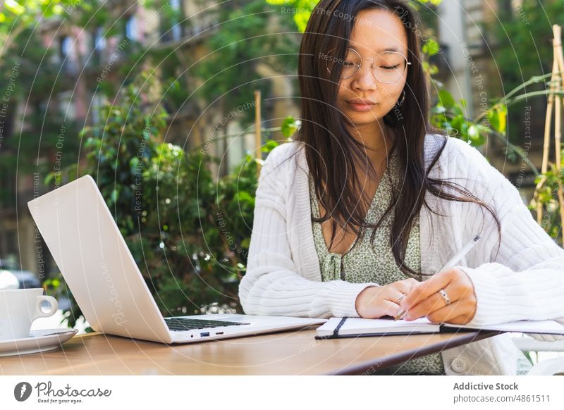 Asiatische selbständige Frau schreibt in einem Café im Freien in ein Notizbuch zur Kenntnis nehmen Notebook schreiben freiberuflich Plan Zeitplan Terrasse