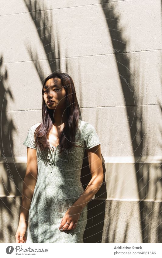 Nachdenkliche asiatische Frau in der Nähe von Gebäuden mit Schatten Wand Stil Sonnenlicht Straße urban Sommer charmant feminin Outfit ethnisch Großstadt Kleid