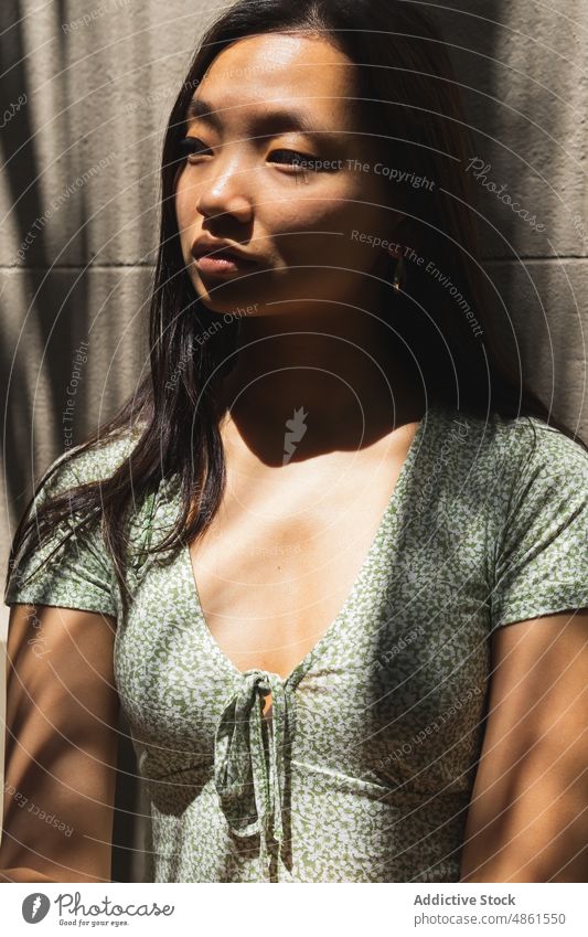Nachdenkliche asiatische Frau in der Nähe von Gebäuden mit Schatten Wand Stil Sonnenlicht Straße urban Sommer charmant feminin Outfit ethnisch Großstadt Kleid