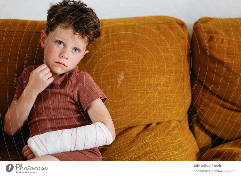 Niedlicher Junge mit gebrochenem Arm auf der Couch ruhen Sofa heimwärts Verletzung Wochenende Arme Wohnzimmer Porträt bandagieren verputzen Gesicht berühren