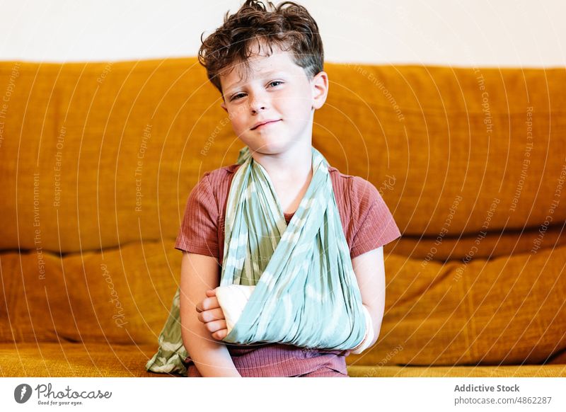 Niedlicher Junge mit gebrochenem Arm auf der Couch ruhen Sofa heimwärts Babytragetuch Verletzung Wochenende Arme Wohnzimmer Porträt bandagieren verputzen Kind