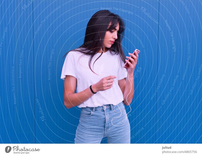 Junges Mädchen arabischer Abstammung benutzt ihr Handy vor einer blauen Wand Ethnizität Spanien jung Frau Zwanziger Jahre trendy Gebäudeaußenseite junge Frau