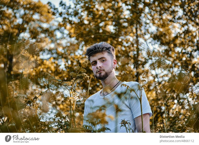 Porträt eines modischen und geheimnisvollen blonden jungen Mannes mit blauen Augen, versteckt hinter einigen Bäumen im Sonnenlicht nachdenklich Selbstliebe
