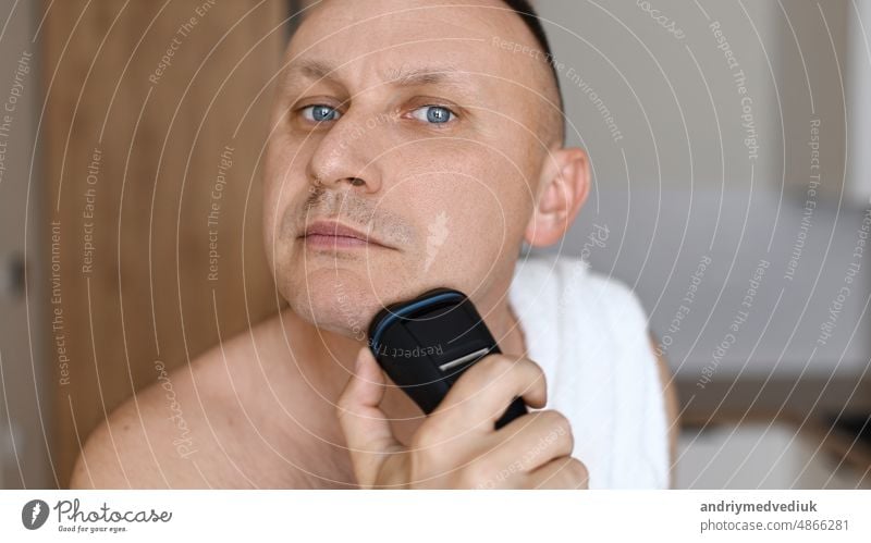 Tägliche Routine männliche Selbstfürsorge. Gutaussehender junger unrasierter Mann mit Handtuch auf den Schultern, der sich mit einem modernen elektrischen Rasierapparat rasiert, während er in die Kamera schaut, drinnen.