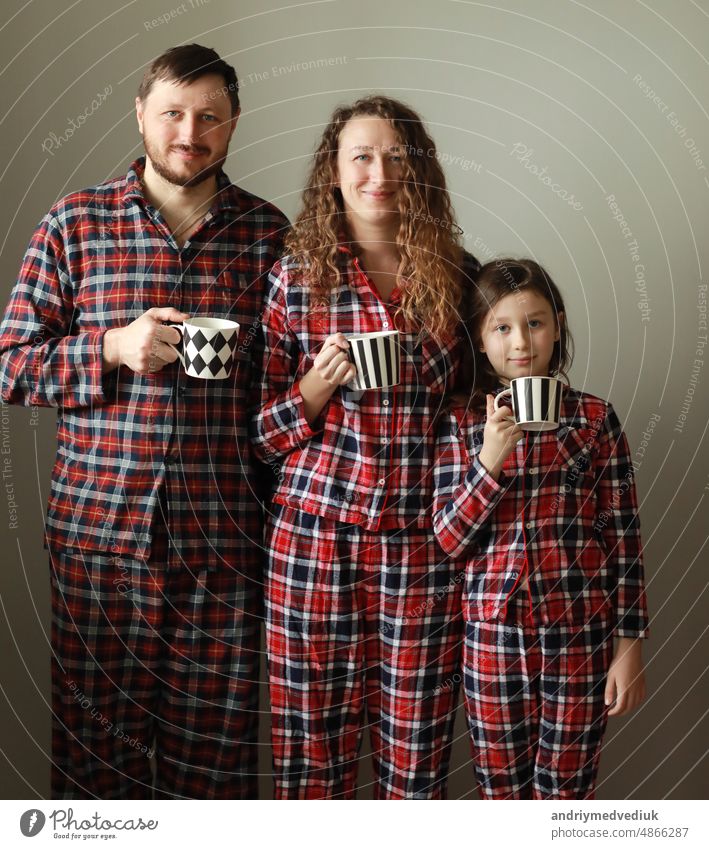Familie im Pyjama auf weißem Hintergrund. Papa, Mama und kleine Tochter mit Tassen Tee. Sie lächeln, albern herum, machen lustige Gesichter und schauen in die Kamera. Vatertag, Muttertag und Babytag.