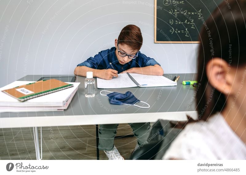 Junge schreibt in der Schule in sein Notizbuch Kind Schüler Klassenraum Maske auf dem Schreibtisch covid-19 Sicherheit schreibend Handdesinfektionsmittel Virus