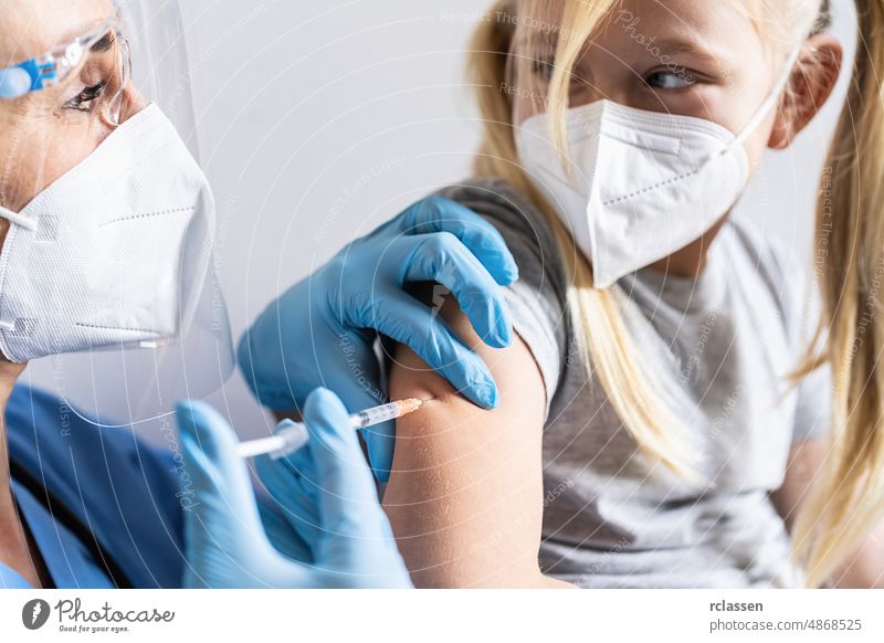 Kleines Mädchen mit Gesichtsmaske in der Arztpraxis wird geimpft. Spritze mit Impfstoff gegen Covid-19 Coronavirus, Grippe, gefährliche Infektionskrankheiten. Injektion nach klinischen Studien für Mensch, Kind.