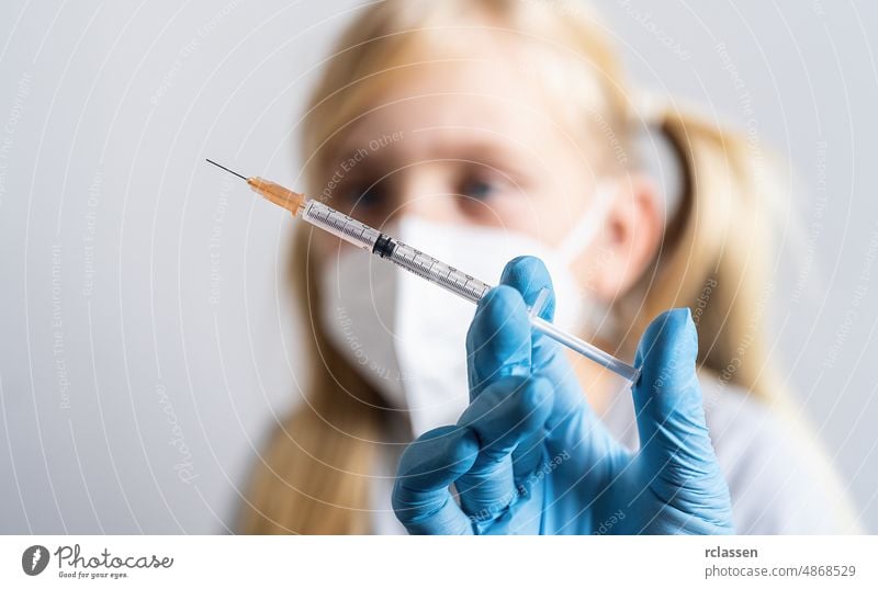 Nahaufnahme einer medizinischen Spritze in der Hand eines Arztes mit einem Impfstoff gegen das Covid-19-Coronavirus, Grippe, und einem blonden Kind im Hintergrund
