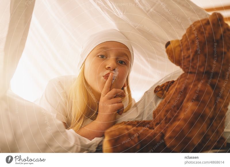 Süßes kleines Mädchen mit einem Teddybär hält den Finger auf die Lippen und zeigt, dass sie schweigen, um ein Geheimnis zu bewahren, während sie auf ihrem Bett in einem Zelt mit Nachtmütze liegt.