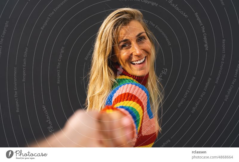 Glücklich aufgeregt Frau zieht Kerl die Hand tragen bunte Regenbogen-Pullover über schwarzem Hintergrund stehen, lgbt feiern Konzeptbild Erwachsener attraktiv