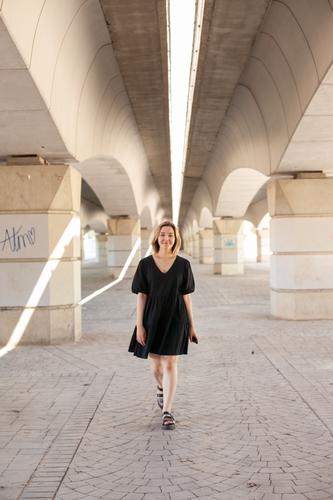 Junge Frau in lässigen schwarzen Kleid zu Fuß unter der Brücke in städtischen Lage in der Stadt. Junge Generation Z Stil Fotoshooting. Echte Menschen Lebensstil in der Stadt. Geometrie in der Stadtarchitektur. Frau zu Fuß in Richtung der Kamera.