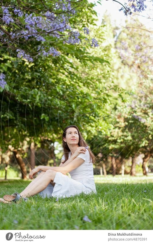 Junge Frau brunette sitzt im Park auf dem grünen Gras im Sommer. Lifestyle offenes Foto im Freien von Frau in Freizeitkleidung gekleidet. Entspannende Momente im Park im Frühling.