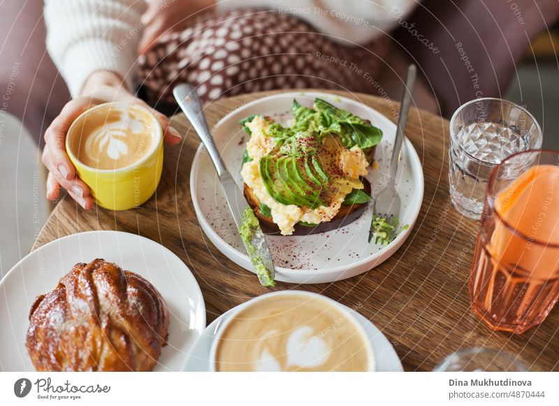 Avocado-Toast auf Holztisch mit Gebäck, Glas Wasser und Kaffeetasse. Cappuccino trinken beim Brunch mit Freunden. Unvoreingenommener Lebensstil mit Essen und Trinken. Millennial Lebensweise. Blick von oben auf den Tisch mit Tellern und Speisen.