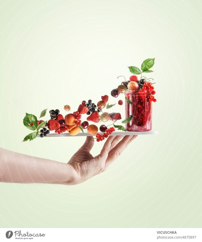 Frauenhand hält Tablett mit rotem Marmeladenglas und verschiedenen fliegenden Beeren und grünen Blättern Hand Beteiligung Glas grüne Blätter blass Hintergrund