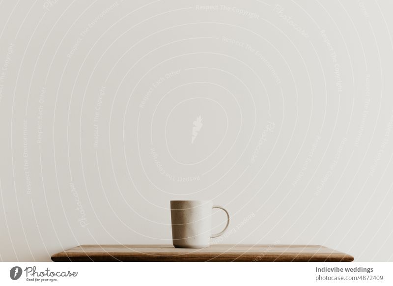 Ein beiger Keramikbecher auf einem braunen Holzbrett vor einer weißen Wand. Handgemachte Kaffeetasse mit Kopierraum. Minimales Konzept, einfaches Design. Tasse